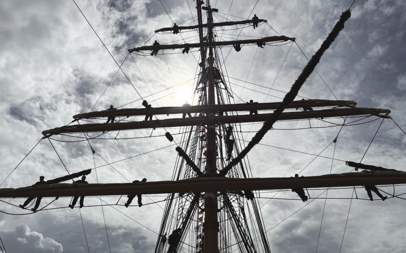 Younger crew members of the USCGC Eagle stand along the spars of the ships mainmast as part of a training exercise.